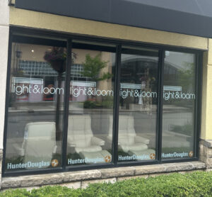 Storefront window with "light & loom" and "HunterDouglas" logos, showing white chairs inside and street reflections outside.